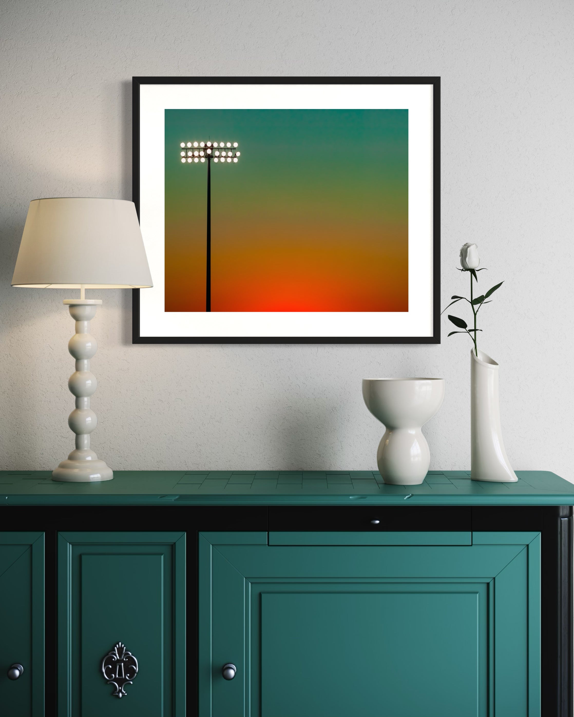 photo of sunset at football stadium single pole of lights left of center, gradient sky teal blue to pale yellow to orange gradient top to bottom in black frame with white matte on white wall above teal furniture piece