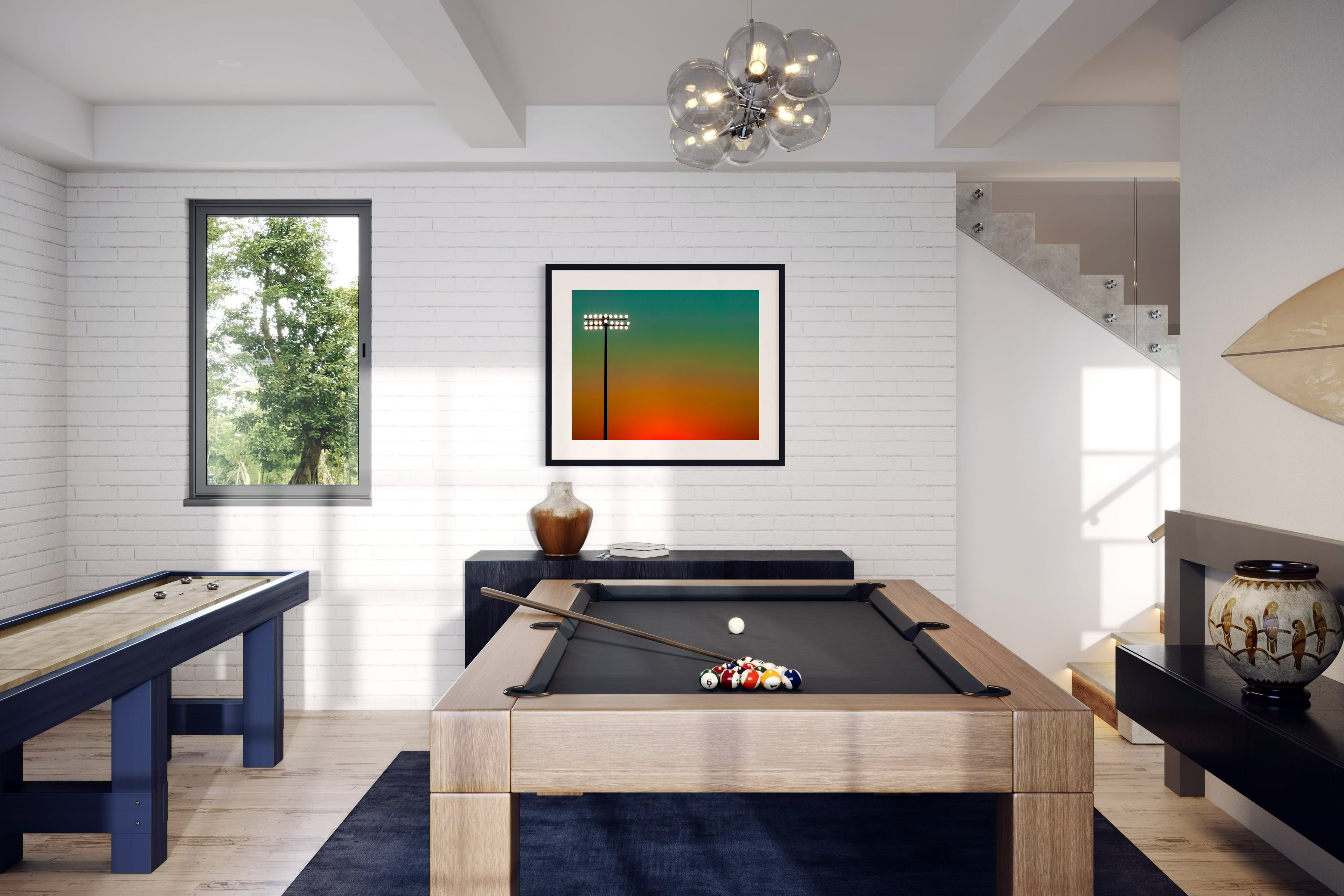 photo of sunset at football stadium single pole of lights left of center, gradient sky teal blue to pale yellow to orange gradient top to bottom in black frame with white matte on white brick wall in game room with pool and shuffle board tables