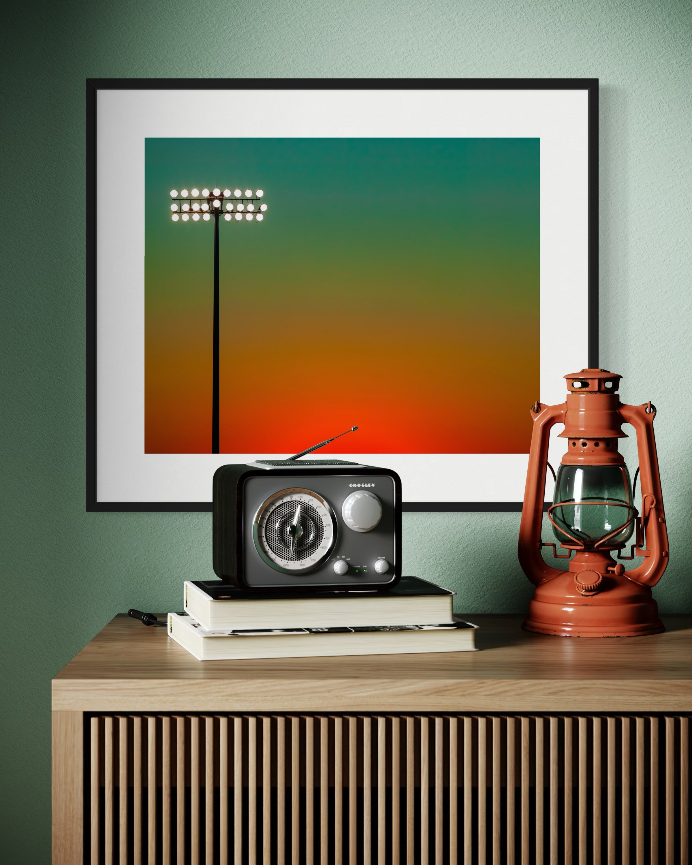 photo of sunset at football stadium single pole of lights left of center, gradient sky teal blue to pale yellow to orange gradient top to bottom in black frame with white matte on light teal wall above modern wooden furniture piece with old radio and lantern on top