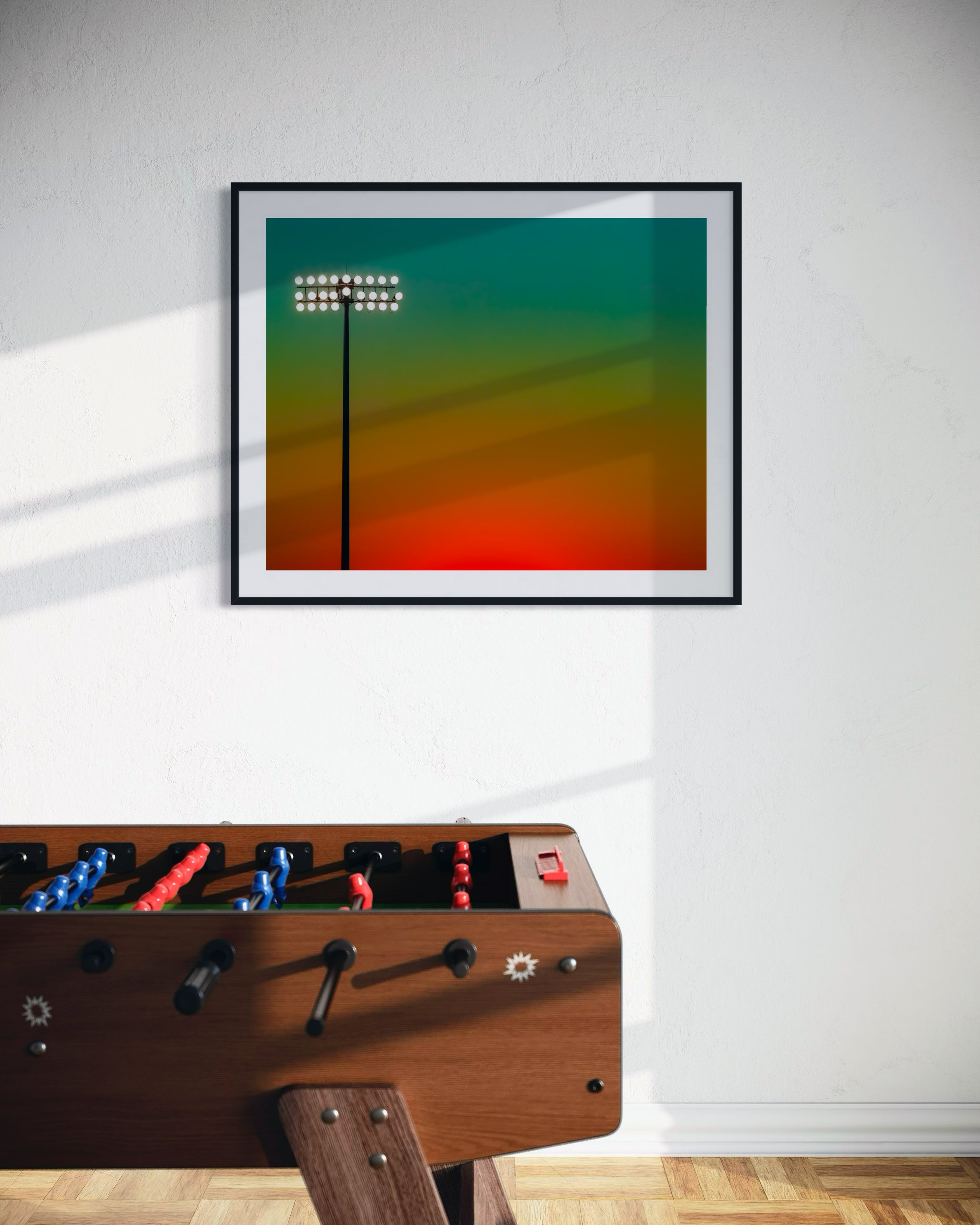 photo of sunset at football stadium single pole of lights left of center, gradient sky teal blue to pale yellow to orange gradient top to bottom in black frame with white matte on white wall in game room foosball table