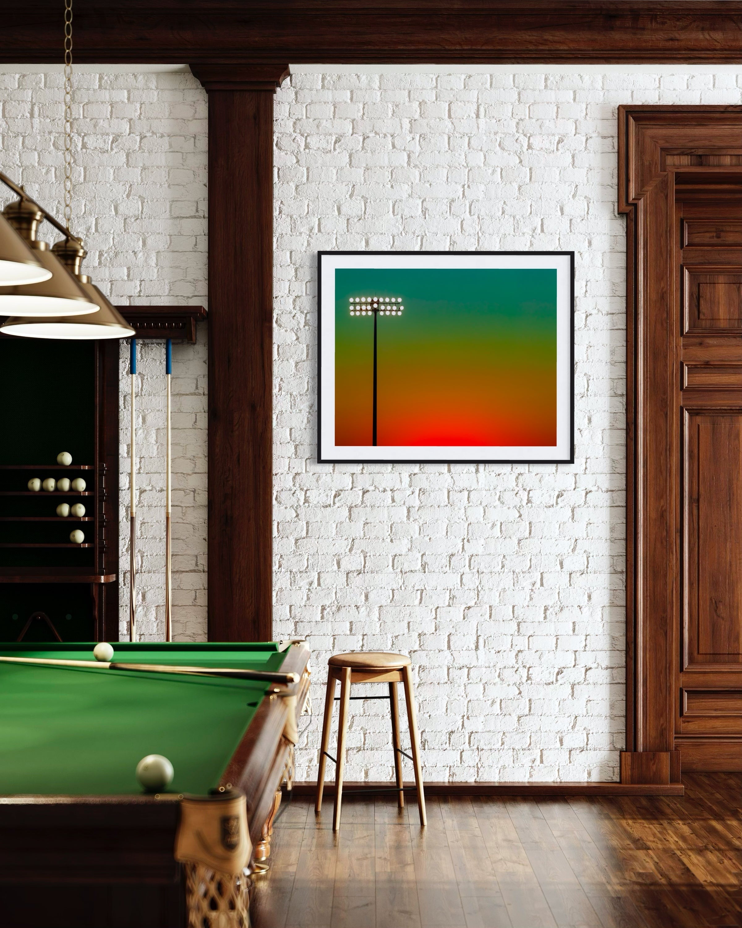 photo of sunset at football stadium single pole of lights left of center, gradient sky teal blue to pale yellow to orange gradient top to bottom in black frame with white matte on white brick wall in game room with pool table