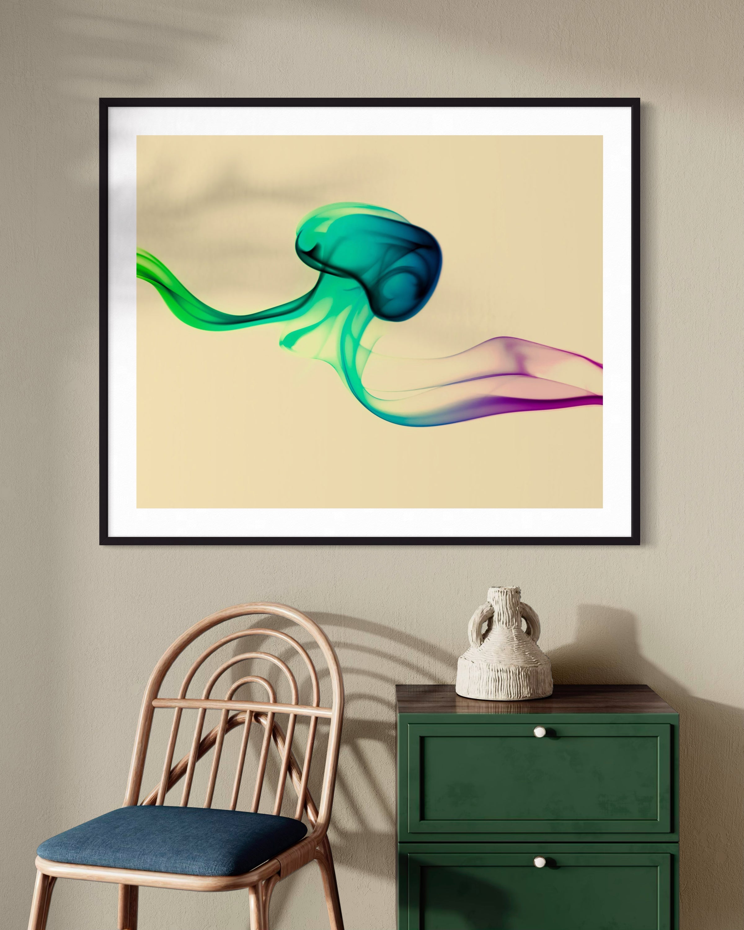 Framed abstract art of a green and purple smoke-like design on a beige wall, with a chair and cabinet below.