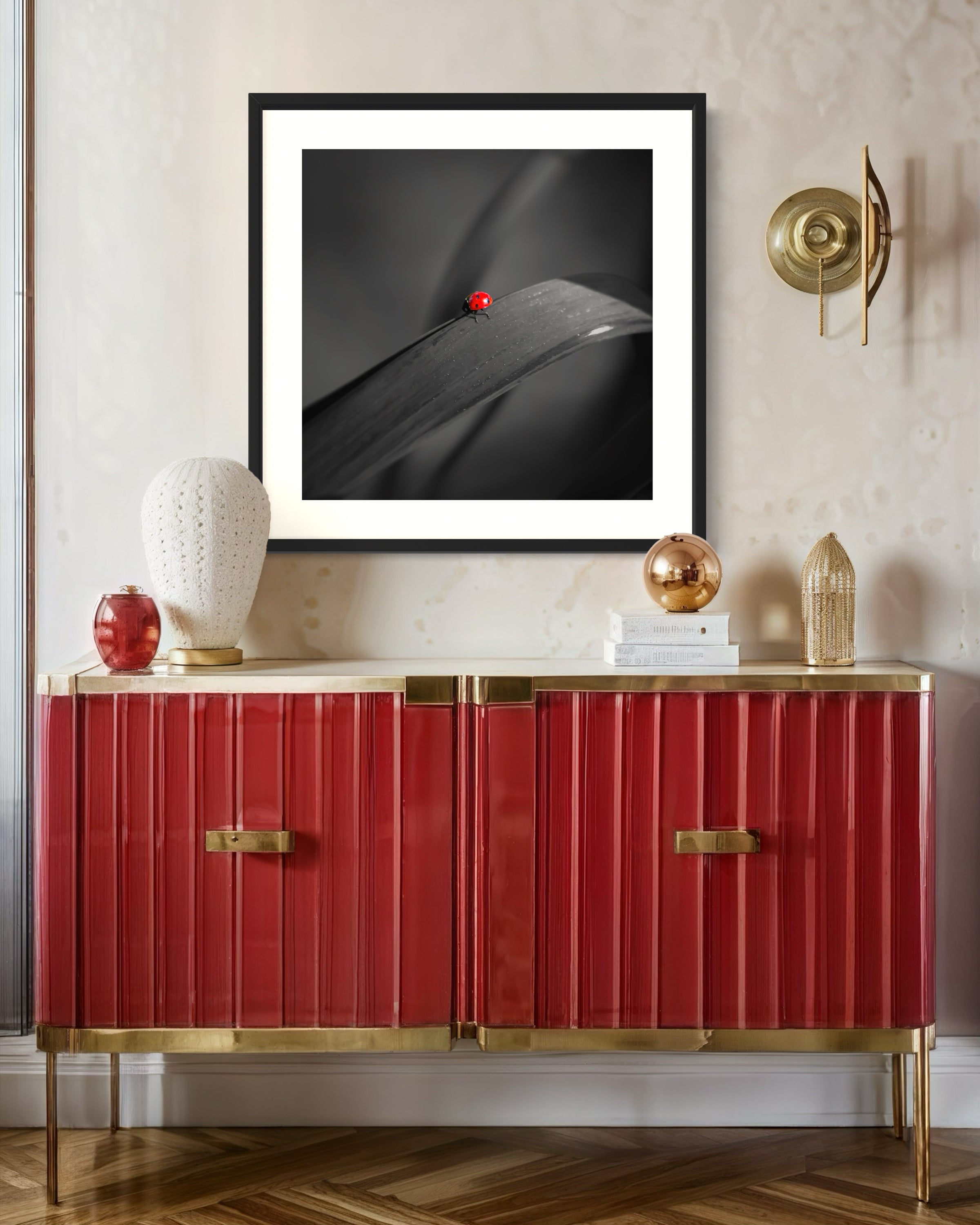 photo of Ladybug in middle of square image, everything except the Ladybug is black and white in black frame on white wall with red table below on hardwood floor