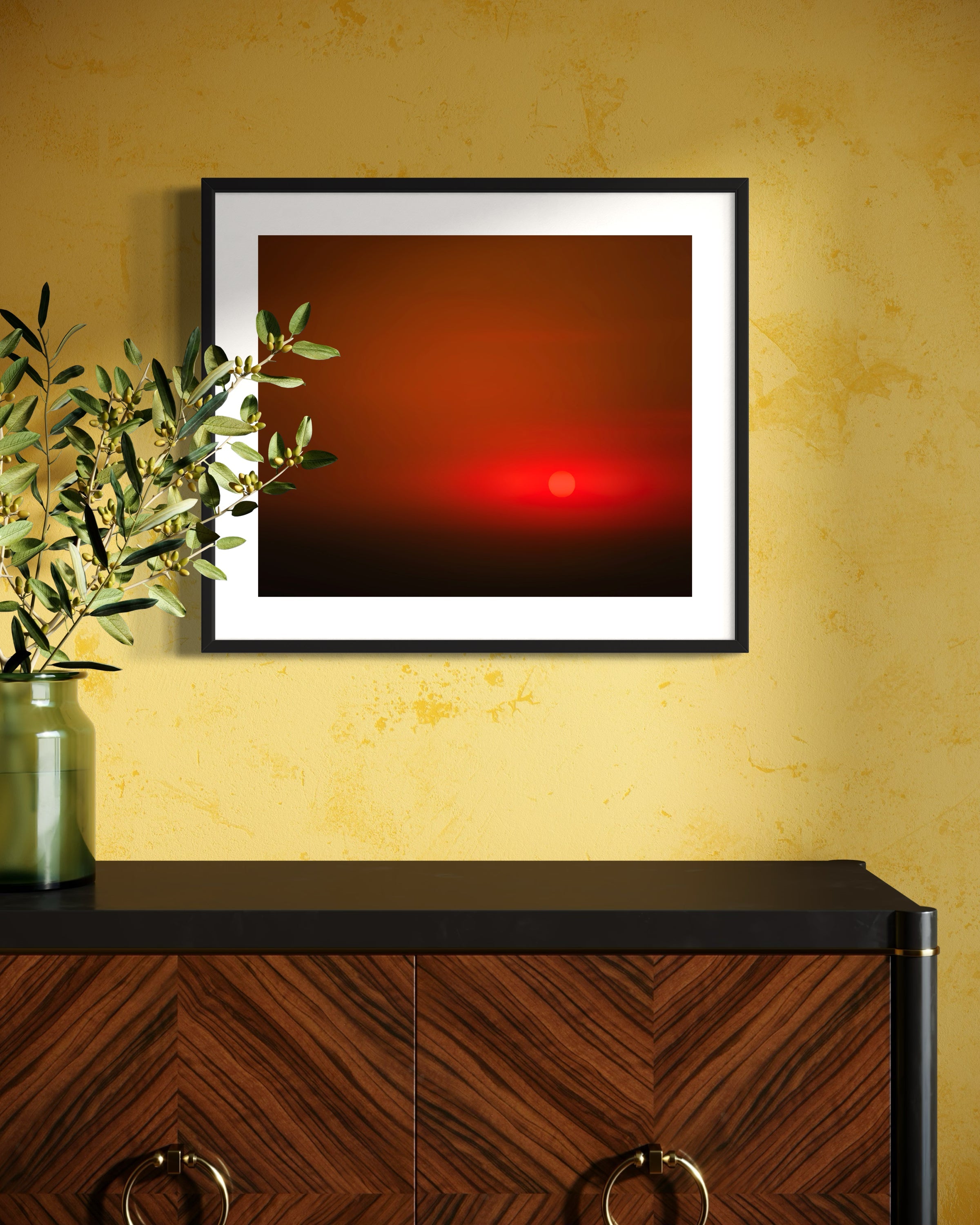 Framed abstract artwork with a red center on a yellow wall above a wooden cabinet.